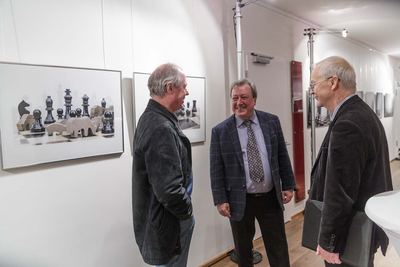 Drei Herren unterhalten sich lachend in der Ausstellung, an der Wand eine Fotografie mit Schachfiguren und Holztieren auf dem Brett.