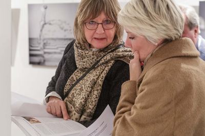 Zwei Besucherinnen blättern gemeinsam im Ausstellungskatalog, hinter ihnen eine Fotografie mit Wasserspritzer über Schachbrettmuster.