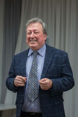 Ein lachender Mann im karierten Sakko mit einer Krawatte voller kleiner Schachfiguren spricht zu den Gästen.