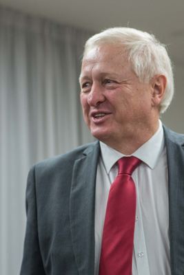 Porträt eines weißhaarigen Mannes in grauem Anzug und roter Krawatte, der lächelnd zur Seite blickt.