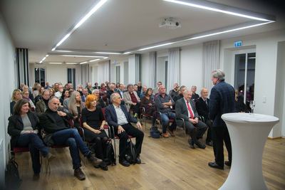 Voll besetzter Saal bei der Vernissage: Das Publikum hört einem Mann zu, der neben einem weiß verhüllten Stehpult steht.