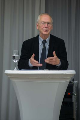 Ein Redner mit Brille spricht gestikulierend an einem weiß verhüllten Stehpult, darauf ein Wasserglas und Manuskriptblätter.