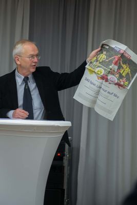 Der Redner am Stehpult hält während seiner Eröffnungsrede eine Zeitungsanzeige zu einem Fußball-Topspiel in die Höhe.