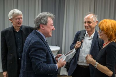 Gespräch nach der Vernissage: Ein Mann im karierten Sakko hält lachend einen Umschlag, umringt von drei Gästen.