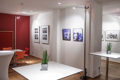 Ausstellungsraum mit Stehtischen und gerahmten Schachfotografien in Schwarzweiß und Blau an weißen Wänden neben einer roten Wand.