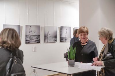 Besucherinnen an einem Stehtisch vor einer Reihe von Fotografien, die Wasserspritzer wie Figuren über einem Schachbrettboden zeigen.