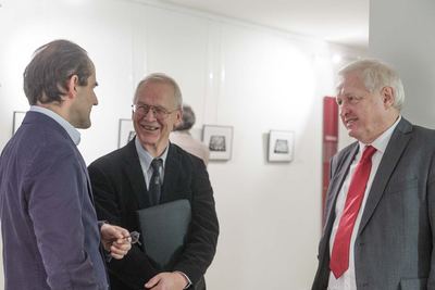 Drei Männer im Gespräch bei der Vernissage, im Hintergrund kleine gerahmte Schachbrett-Grafiken an der Wand.