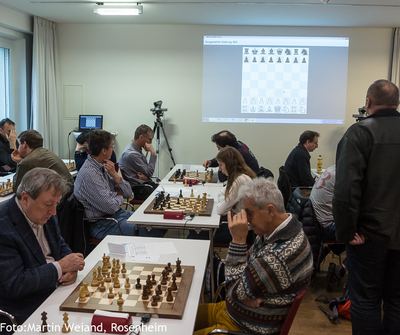 Partien im Turniersaal, dahinter Kamera auf Stativ und Leinwand mit der ausgewählten Schach960-Grundstellung