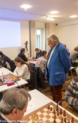 Zuschauer im blauen Sakko steht zwischen den Tischreihen und verfolgt die laufenden Partien des Schach960-Turniers