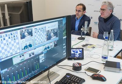 Kommentatorentisch der Liveübertragung: Bildmischer-Monitor mit Brettern und Spielern, dahinter zwei Kommentatoren vor Sponsorenwand