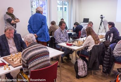 Blick in den Turniersaal mit mehreren Brettern, stehenden Zuschauern und Kamera auf Stativ am Fenster