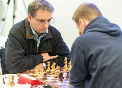 Turnierspieler mit Brille konzentriert über dem Brett, die Hand am Tisch, Gegner im Kapuzenpullover im Vordergrund
