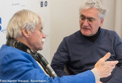 Zwei ältere Herren im lebhaften Gespräch vor der Sponsorenwand, einer gestikuliert mit der Hand