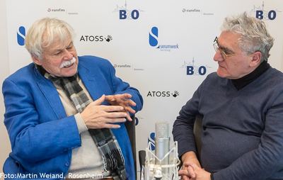 Zwei Gesprächspartner sitzen am Studiomikrofon vor der Logowand, einer erklärt mit beiden Händen