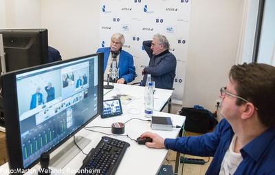 Techniker am Bildmischpult verfolgt auf dem Monitor das Studiogespräch, das im Hintergrund vor der Logowand stattfindet
