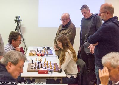 Zuschauer stehen dicht hinter einer Spielerin und schauen auf ihr Brett, links weitere Partien und eine Kamera