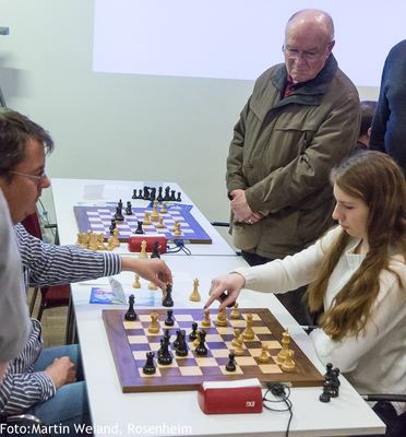 Spielerin und Spieler ziehen gleichzeitig am Brett, ein älterer Zuschauer verfolgt die Partie