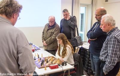 Mehrere Zuschauer umringen einen Turniertisch, an dem eine junge Spielerin mit Schwarz am Zug überlegt