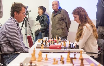 Lange Tischreihe mit Partien, Zuschauer stehen an der Wand, vorn eine Spielerin und ihr Gegner im Profil