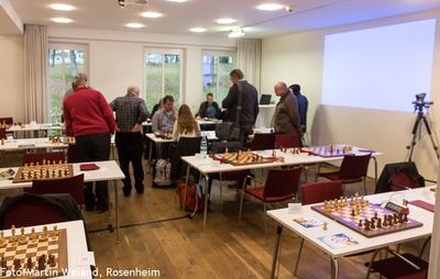 Spielsaal mit Fenstern zum Park, Spieler und Zuschauer zwischen den Brettern, rechts Leinwand und Kamera