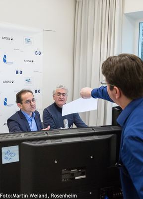Regie hinter dem Monitor: ein Mann reicht ein Blatt zu den Kommentatoren am Mikrofon vor der Logowand