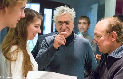 Teilnehmer beugen sich lächelnd über ein Blatt Papier und analysieren gemeinsam, im Hintergrund weitere Zuschauer