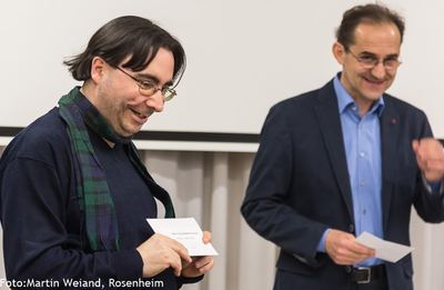 Preisträger mit Schal blickt lächelnd auf seinen Umschlag, daneben der Überreicher mit weiteren Umschlägen