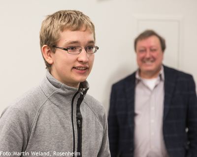 Junger Turnierteilnehmer mit Brille im grauen Pullover spricht bei der Preisverleihung, dahinter ein lächelnder Mann