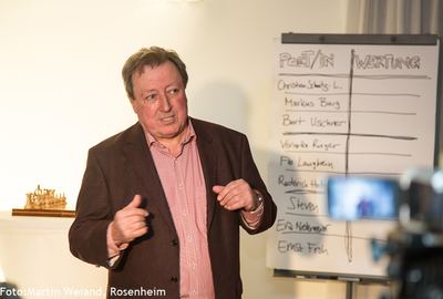 Ein Mann im Sakko spricht vor einem Flipchart mit der Liste der Slam-Teilnehmer und der Spalte „Wertung“; links ein Schachspiel.