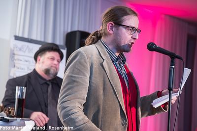 Ein Slam-Poet im Cordsakko trägt am Mikrofon einen Text vom Blatt vor; im Hintergrund der Moderator mit Hut vor dem Flipchart.