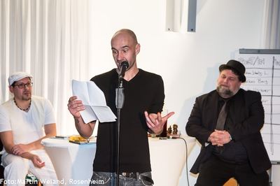 Ein Slam-Poet in schwarzem Shirt trägt gestikulierend vom Blatt vor, flankiert von den beiden sitzenden Moderatoren.