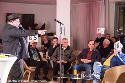 Publikumswertung beim Poetry Slam: Zuschauer halten Punktetafeln mit 8,5, 7,7 und 8,8 hoch, der Moderator zeigt auf sie.