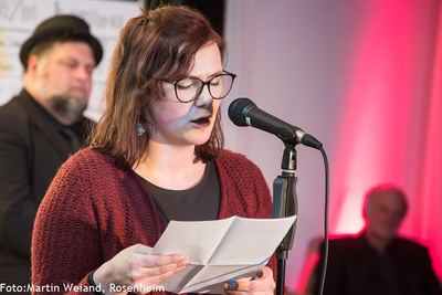 Eine Slam-Poetin mit Brille liest am Mikrofon von einem Blatt; hinter ihr der Moderator mit Hut.