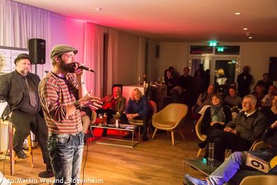 Ein Slam-Poet mit Schiebermütze und Ringelpulli performt frei am Mikrofon vor dem dicht besetzten, rot beleuchteten Saal.