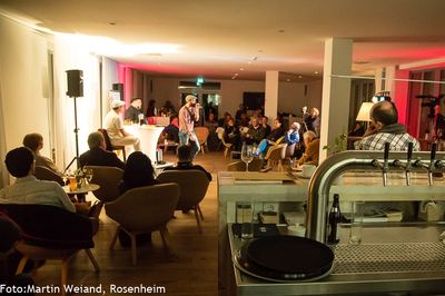 Blick von der Bar über den vollen Saal mit Sesseln zur kleinen Bühne, auf der ein Slam-Poet am Mikrofon steht.