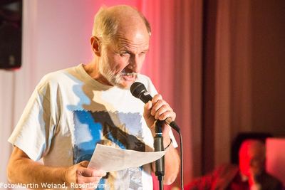Ein älterer Slam-Poet mit weißem Bart hält das Mikrofon und liest im roten Bühnenlicht von einem Blatt vor.