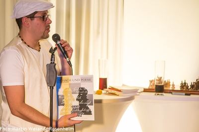 Ein Moderator mit weißer Mütze hält den Ausstellungskatalog „Schach und Poesie“ in die Höhe; dahinter Schachfiguren auf dem Tisch.