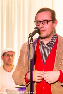 Ein Mann mit Brille im Cordsakko und roter Weste spricht mit gefalteten Händen ins Mikrofon; im Hintergrund ein Moderator.
