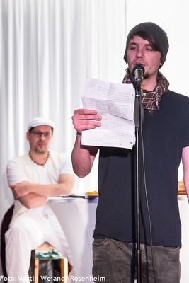 Ein junger Slam-Poet mit Mütze und Schal liest von einem handbeschriebenen Zettel; dahinter sitzt ein Moderator mit verschränkten Armen.