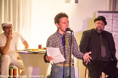 Ein Slam-Poet im karierten Hemd trägt mit ausgebreiteter Hand vor, links und rechts sitzen die beiden Moderatoren.