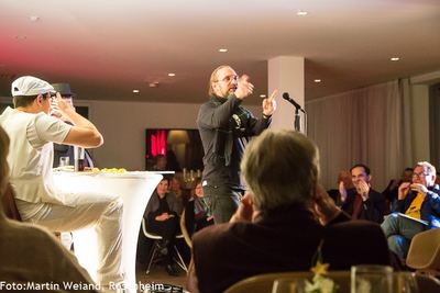 Ein Slam-Poet mit Brille und Schal steht gestikulierend vor dem Mikrofon, während das Publikum im Saal applaudiert.