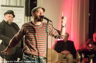 Ein bärtiger Slam-Poet mit Schiebermütze performt mit geschlossenen Augen und erhobener Hand am Mikrofon.