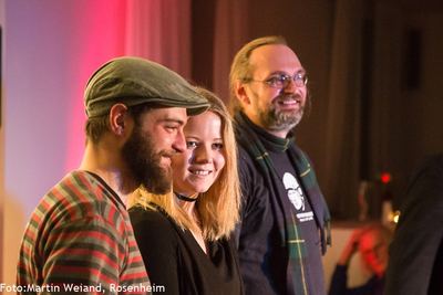 Drei Finalisten des Poetry Slams stehen lächelnd nebeneinander vor rot beleuchtetem Vorhang.
