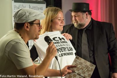 Siegerehrung: Die Gewinnerin hält ein T-Shirt mit der Aufschrift „Poetry Slam gewonnen“, daneben eine Urkunde „Schach und Poesie“.
