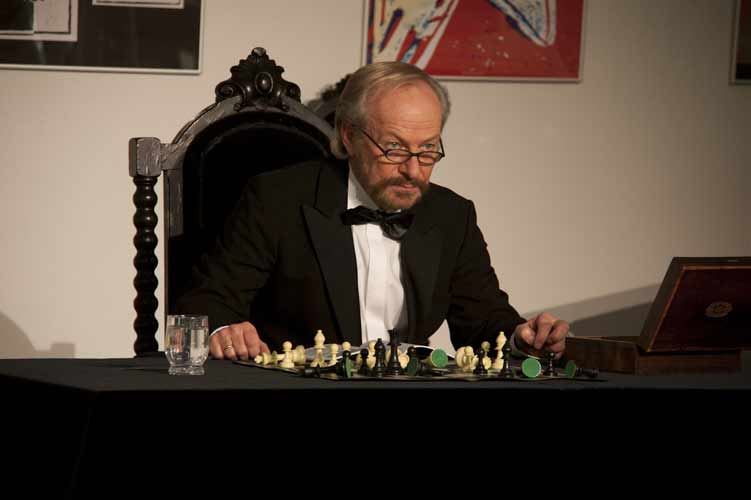 Ein Schauspieler in Smoking, Fliege und Brille sitzt auf einem geschnitzten Stuhl vor einem Schachbrett mit Figuren