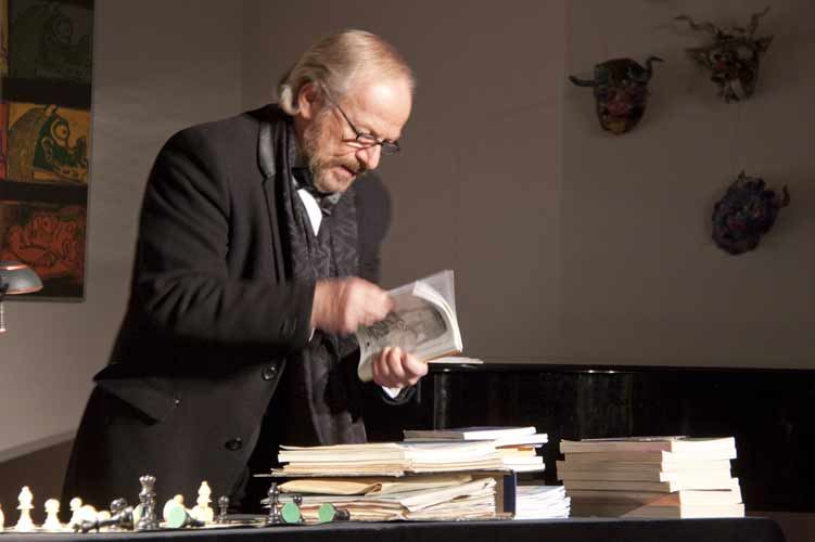 Der Schauspieler im Gehrock blättert in einem alten Heft, vor ihm Papierstapel, Bücher und Schachfiguren auf dem Tisch