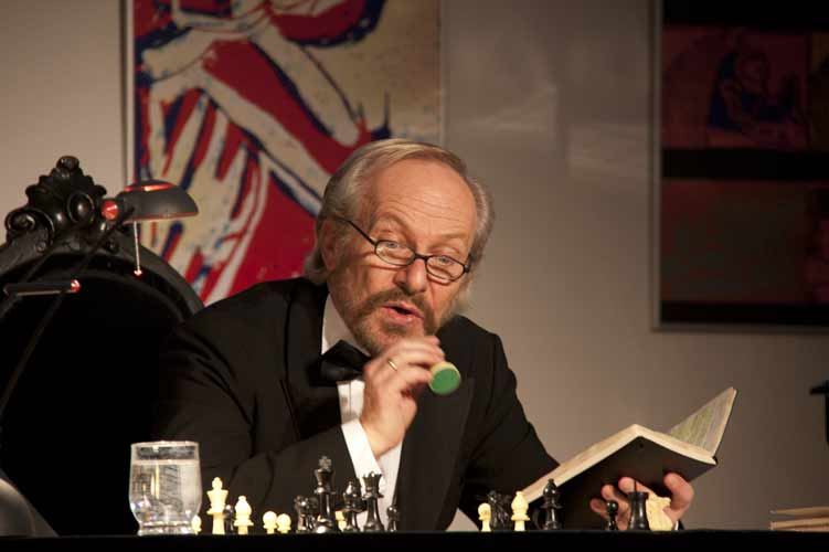 Der Schauspieler hält mit aufgeschlagenem Notizbuch eine Schachfigur hoch und spricht zum Publikum