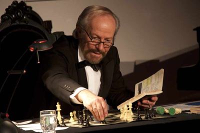 Mit einem aufgeschlagenen Schachheft in der Hand zieht der Schauspieler eine Figur auf dem Brett