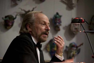 Der Schauspieler im Smoking spricht in der Diskussion, im Hintergrund unscharf bunte Masken an der Wand