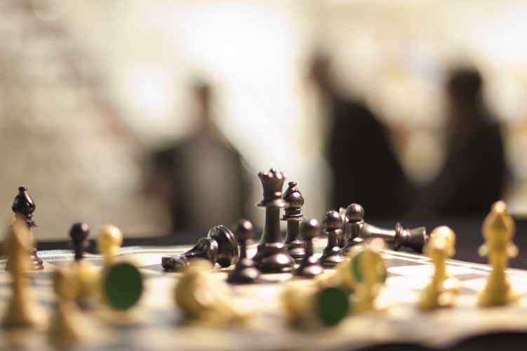 Schachbrett mitten in der Partie aus Spielerperspektive, schwarze und helle Figuren, im unscharfen Hintergrund dunkle Silhouetten